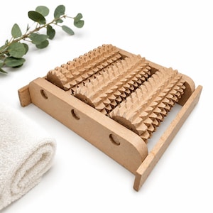 Op de afbeelding: Een lichtbruine houten voetmassager met vier parallelle rollers met een stekelig, getextureerd oppervlak. De rollers zijn gemonteerd in een rechthoekig houten frame met drie cirkelvormige uitsparingen aan de voorzijde voor handmatig gebruik.