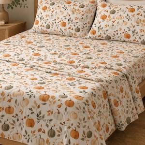 Peut inclure: Un ensemble de draps de lit et deux taies d'oreiller avec un motif répétitif de citrouilles orange et vert sourd entremêlées de feuilles brunes et vertes sur un fond blanc, recouvrant un lit avec une tête de lit et un cadre en bois.
