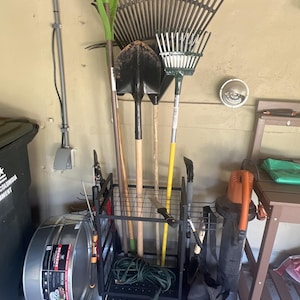 May include: A black metal garden tool organizer rack holding various long-handled tools including a green leaf rake, a black shovel, and a yellow-handled rake. The text ZorevaCraft is visible at the top and bottom. A galvanized metal tub sits to the left.