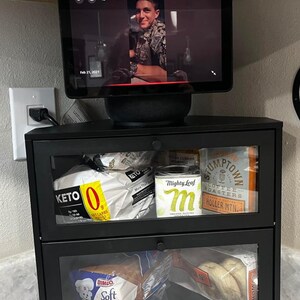 May include: A black two-tier storage cabinet with clear glass doors, containing various food items including Keto bread, Mighty Leaf tea, Stumptown coffee, and Bimbo bread. A smart display device with a screen showing a person is placed on top.