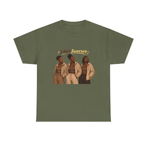 Może przedstawiać: Oliwkowozielony t-shirt z krótkim rękawem z nadrukiem graficznym przedstawiającym trzy osoby w beżowych kurtkach i ubraniach w neutralnych odcieniach. Nad postaciami znajduje się napis I Aint Insecure stylizowaną złotą czcionką z gwiazdkami.