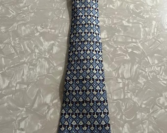 May include: A necktie featuring a repeating geometric pattern in shades of light blue, dark blue, and cream, accented with small dark red circular details. The fabric displays a structured, symmetrical design with interlocking shapes throughout the length.