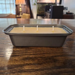 Puede incluir: Un molde rectangular de metal gris lleno de cera lisa de color beige claro. La vela presenta tres mechas blancas espaciadas uniformemente que sobresalen de la superficie. El recipiente tiene un borde ancho y plano con lados ligeramente inclinados.