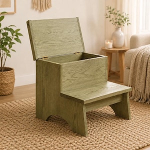 Puede incluir: Un taburete de madera color verde salvia con una tapa abatible que se abre para revelar un compartimento de almacenamiento. El taburete presenta un diseño de dos niveles con un escalón inferior y una sección trasera más alta.