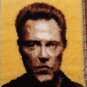 May include: A close-up of a rug hooking portrait of a man with brown hair and a serious expression. The man is depicted against a yellow background.