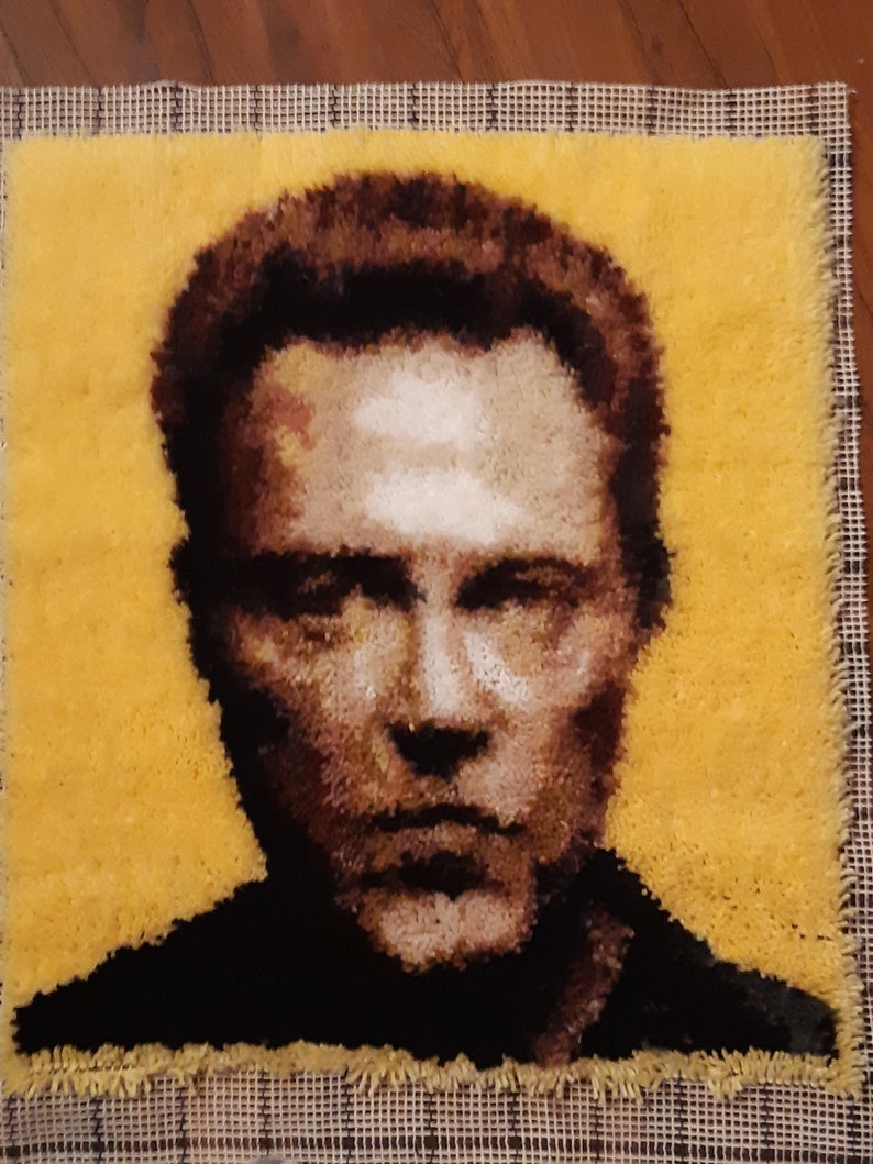 May include: A close-up of a rug hooking portrait of a man with brown hair and a serious expression. The man is depicted against a yellow background.