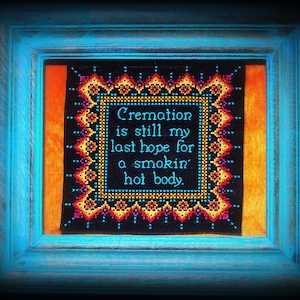 May include: A framed cross-stitch with a black background and colourful border. The text reads "Cremation is still my last hope for a smokin' hot body."