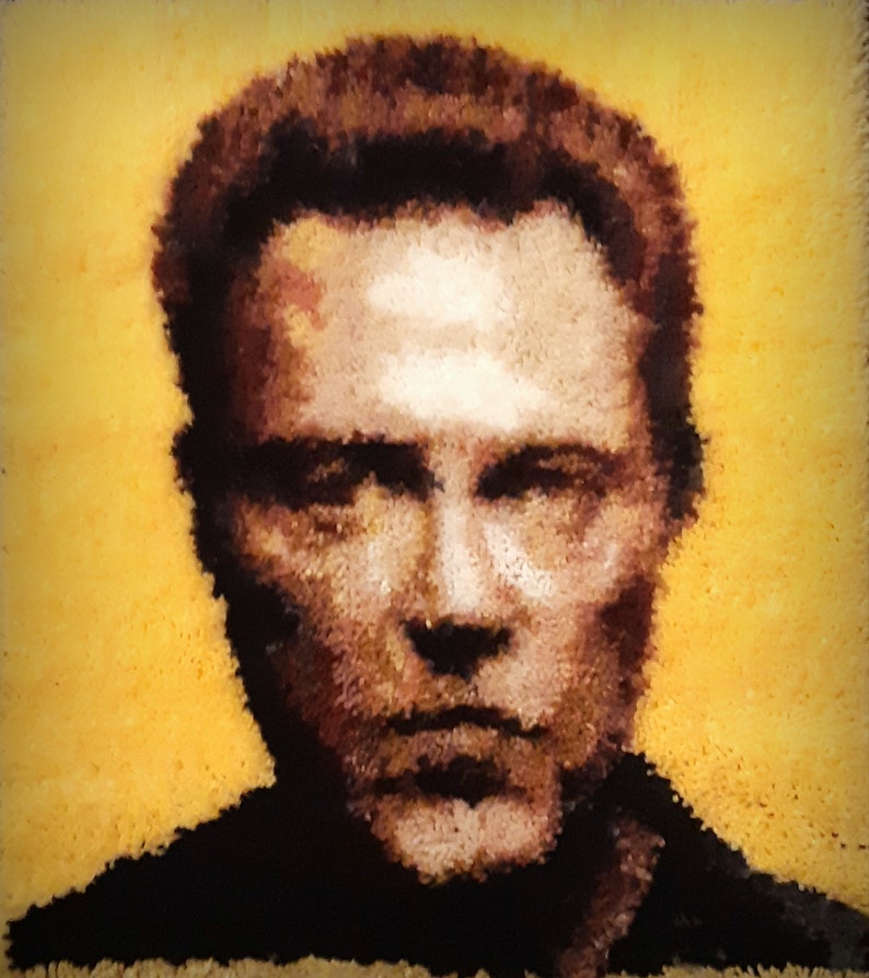 May include: A close-up portrait of a man with short brown hair. The image is rendered in a pixelated style, resembling a rug or tapestry. The background is a solid yellow color.