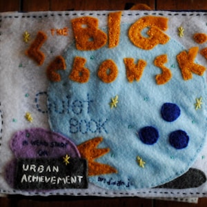 Può includere: Un libro silenzioso in feltro blu e bianco con il testo "The Big Ebowsk" in lettere di feltro arancioni. Il libro ha una toppa in feltro viola con il testo "A Weird Story on Urban Achievement".
