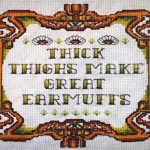 May include: Cross-stitch embroidery with a white background and a brown and green frame. The embroidery says "Thick Thighs Make Great Earmuffs".
