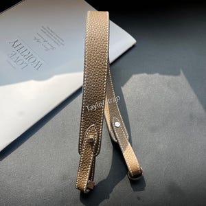 May include: A taupe-colored textured leather bag strap featuring white contrast stitching along the edges. The strap includes silver-toned metal hardware, specifically a small stud and a circular ring attachment, with the text TaylorStrap visible.