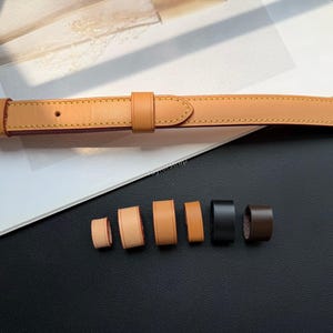 May include: A tan leather belt strap with a gold-toned metal buckle and a matching sliding keeper loop. Below the belt are six additional leather loops in varying widths and colors, including tan, light brown, black, and dark brown shades.