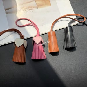 May include: Four leather tassels with loops, featuring a brown one with a light grey top, a pink one with a light pink top, a solid tan one, and a solid black one. Each tassel has a fringe design and a thin strap for attachment.