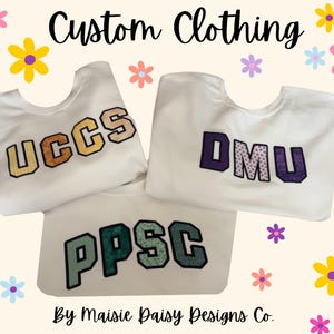 May include: Three white crewneck sweatshirts featuring large varsity-style letters. One displays UCCS in yellow and brown, another DMU in purple and white, and the third PPSC in green and black. The text Custom Clothing and By Maisie Daisy Designs Co. appears.