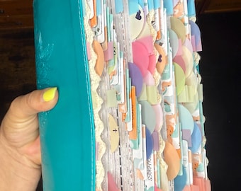 May include: A teal-colored book with a smooth, glossy cover and a floral embossed design. The pages are filled with numerous colorful, semi-circular paper tabs in shades of pink, blue, orange, and green, accented with cream-colored lace trim.