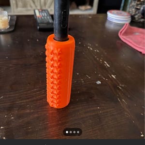 May include: A bright orange TIG welding torch handle with a textured, knurled grip surface. The handle is cylindrical and attached to a black metal shaft. The design features a repeating geometric pattern along the exterior for enhanced grip.