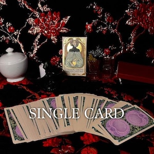 May include: A deck of tarot cards fanned out with a purple and green floral back design, accompanied by a vertical card box featuring an illustration of a person in a gold halo, titled Ethereal Visions Illuminated Tarot Deck by Matt Hughes.