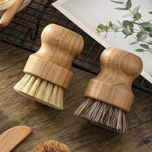 Op de afbeelding: Twee ronde houten afwasborstels met ergonomische handgrepen en stijve haren. De ene borstel heeft lichtgekleurde natuurlijke haren, terwijl de andere donkerdere, grovere haren heeft. Beide borstels hebben gladde, lichtbruine houten bovenkanten.