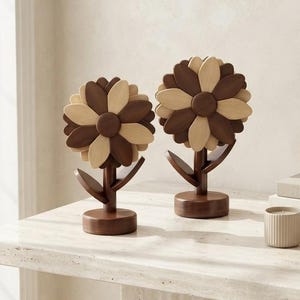 Puede incluir: Dos esculturas decorativas de flores hechas de madera clara y oscura, cada una con un centro circular rodeado de pétalos alternos, montadas sobre una base cilíndrica. Las flores están erguidas y muestran un acabado de veta de madera natural.