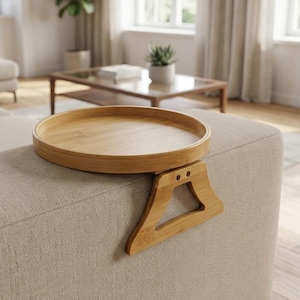 May include: A round, light brown bamboo sofa arm tray with a flat circular surface and a hinged, triangular support bracket designed to grip the side of a couch armrest, providing a stable platform for holding small items or beverages.