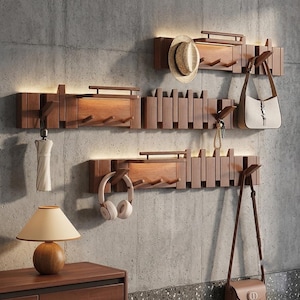Può includere: Tre appendiabiti da parete orizzontali in legno con illuminazione a LED integrata e ganci multipli. I supporti presentano un design a listelli e sostengono un cappello di paglia, una borsa bianca, un ombrello chiuso e cuffie bianche.
