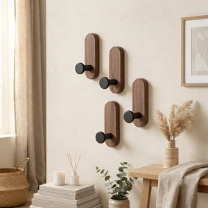 Puede incluir: Cuatro percheros de pared, cada uno con una base vertical de madera marrón oscuro de forma rectangular redondeada y un gancho cilíndrico negro mate que sobresale. Los ganchos están dispuestos en un patrón escalonado sobre una pared clara.