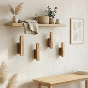 May include: Four vertical light brown wooden wall hooks mounted on a textured off-white wall. Above the hooks, a light brown wooden floating shelf holds a speckled vase with dried grass, a folded beige cloth, a woven basket, and a small plant.