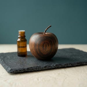 May include: A dark brown wooden apple sculpture with a visible natural grain pattern and a small stem, positioned next to a small amber glass bottle with a screw-top cap. The items are placed on a rectangular black slate board with a rough edge.