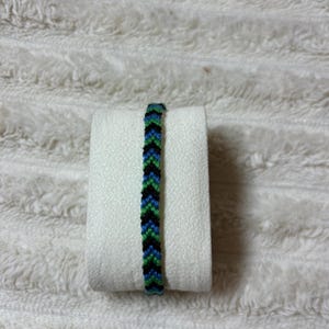 Peut inclure: Un bracelet d'amitié fait main présentant un motif chevron répétitif dans des tons alternés de fil noir, vert vif et bleu roi. Cet accessoire textile tissé affiche un design en forme de V cohérent sur toute sa longueur.