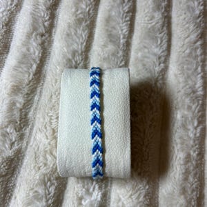 Peut inclure: Un bracelet d'amitié tissé présentant un motif à chevrons répétitif dans des tons de bleu foncé, bleu moyen et blanc. Le bracelet est fabriqué à partir de fil fin et affiche un design en forme de V symétrique et cohérent sur toute sa longueur.
