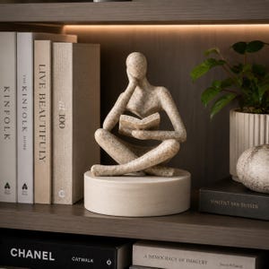 May include: A beige speckled abstract sculpture of a seated figure reading a book, resting on a smooth cylindrical base. The figure has a faceless head resting on one hand, with legs crossed, positioned between several books and a green potted plant.
