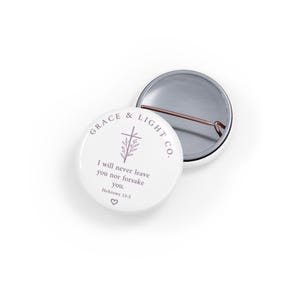 Könnte beinhalten: Ein runder, weißer Ansteckbutton mit dem Text Grace & Light Co. in einem kreisförmigen Bogen, einer floralen Kreuzillustration und dem Bibelvers I will never leave you nor forsake you. Hebrews 13:5 mit einem kleinen Herzsymbol am unteren Rand.