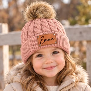 May include: A dusty rose cable-knit beanie featuring a large faux fur pom-pom on top and a folded cuff. A rectangular tan leather patch is attached to the front of the cuff with four silver rivets, displaying the name Emma in black script.