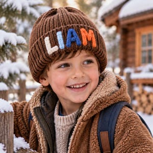 May include: A brown ribbed knit beanie featuring the name LIAM in large, multi-colored yarn letters. The letters are stitched onto the front cuff in white, light blue, dark blue, and orange, creating a textured and bold appearance.