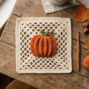 May include: A square off-white crochet granny square featuring a textured orange pumpkin with a green stem in the center. The piece is accompanied by a wooden crochet hook, a small ball of orange yarn, and several dried autumn leaves.