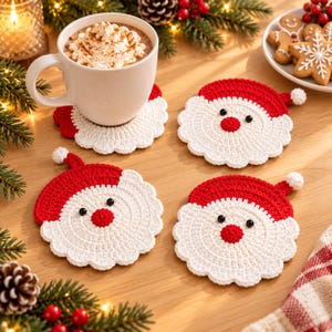 May include: Four round crochet coasters shaped like Santa Claus faces, featuring a red hat with a white pom-pom, a white beard, two black bead eyes, and a red nose. One coaster sits under a white mug filled with whipped cream and cinnamon.