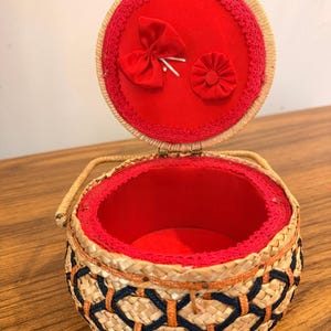 May include: A round woven basket with a hinged lid, featuring a red fabric interior and a red fabric-lined lid decorated with a small red bow and a circular fabric rosette. The exterior is woven with tan straw and accented with navy blue and orange cord.
