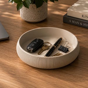 Può includere: Un vassoio portaoggetti rotondo color beige con una texture esterna a coste. L'interno del vassoio contiene una chiave per auto nera, una chiave in metallo dorato, una penna a sfera nera e oro, una clip nera e dei fermagli dorati.