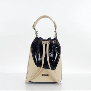 Puede incluir: Un bolso tipo saco con diseño bicolor que combina una sección superior en negro brillante y una sección inferior en beige mate. Incluye un cierre de cordón ajustable, un asa superior con herrajes metálicos y una pequeña placa con logotipo.