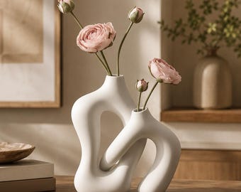 May include: Two interconnected white ceramic vases with smooth, organic, abstract shapes. The taller vase holds two pink ranunculus flowers and one bud, while the shorter vase holds one pink ranunculus flower and one bud, creating a cohesive display.