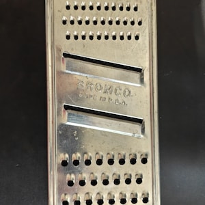 May include: A rectangular silver-colored metal kitchen grater and slicer featuring two horizontal blades and rows of circular perforations. The center of the tool is embossed with the text BROMCO and MADE IN U.S.A. It has a wire handle at each end.