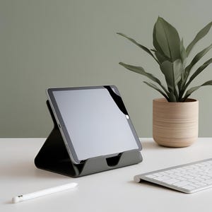 May include: A dark gray tablet stand holding a silver tablet with a black screen. The stand has a triangular profile with a front ledge. A white stylus lies on the surface, and a white wireless keyboard is positioned to the right of the stand.