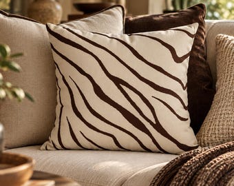 May include: A square throw pillow featuring a cream-colored fabric base with a dark brown zebra stripe pattern. The pillow is positioned among other cushions, including a solid dark brown pillow and a textured beige pillow.