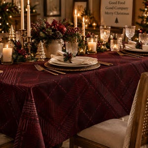 May include: A deep burgundy tablecloth featuring a subtle, repeating geometric herringbone pattern. The fabric drapes over a dining table set with white plates, clear textured glassware, gold cutlery, and cream-colored cloth napkins.