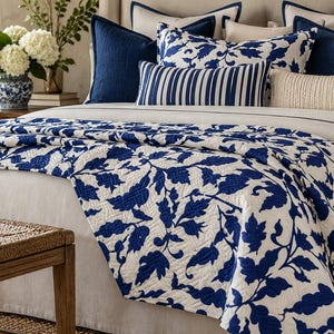 May include: A bed dressed in a white quilt featuring a bold blue botanical leaf pattern. The bedding includes several pillows in solid navy blue, a blue and white striped lumbar pillow, and a textured cream-colored lumbar pillow.