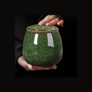 Op de afbeelding: Een ronde keramische pot met een glanzend, diepgroen kristalglazuur met een textuurrijk, gebarsten patroon. De pot heeft een bijpassend deksel met een bruine rand, een glad, reflecterend oppervlak en een donkere, ongeglazuurde onderkant.