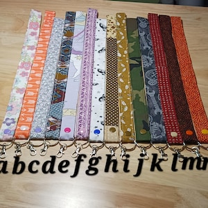 May include: A collection of fourteen fabric lanyards arranged in a row, each featuring a unique pattern including floral, geometric, camouflage, and solid prints. Each lanyard is secured with a snap button and includes a metal swivel lobster clasp.