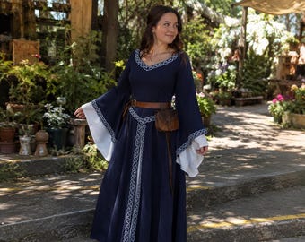 May include: A floor-length navy blue dress featuring long, flared sleeves with white undersleeves, a V-neckline, and white decorative trim along the neckline, sleeves, and center. A brown leather belt with an attached small pouch is worn at the waist.