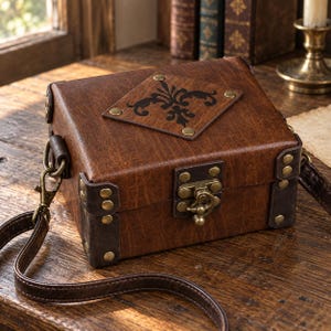 May include: A rectangular brown structured bag with a textured finish, featuring dark brown corner reinforcements secured with brass-toned rivets. A central brass latch closure and a diamond-shaped plaque with a black filigree design are visible.