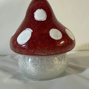 May include: A mushroom-shaped decorative object featuring a glossy red cap covered in fine red glitter and accented with several smooth, circular white spots. The base of the mushroom is rounded and coated in a dense layer of shimmering silver glitter.